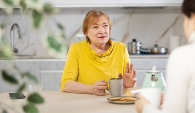Indignant elderly woman with cup of tea sits in kitchen and listens to daughter.Women talk about family difficulties.Concept of troubled family relationships clipart