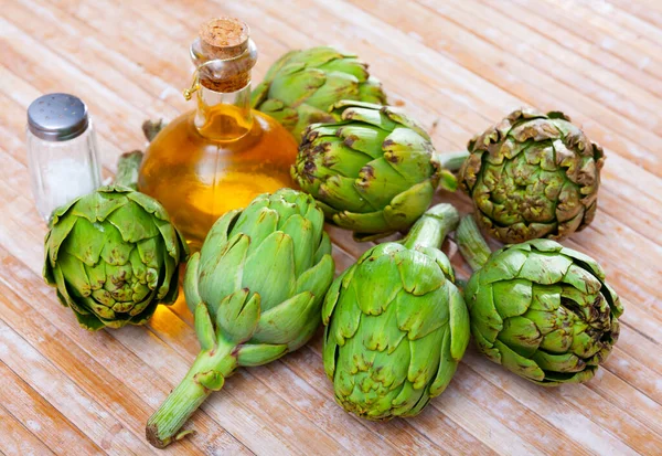 stock image Bunch of artichokes placed over wooden background with saltcellar and glass oil bottle.