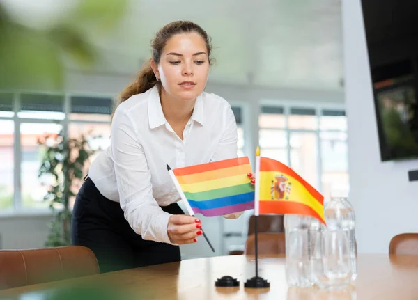 会議室でスペインの国旗と水のボトルの横に小さなLgbtの旗をテーブルの上に置く積極的な若い女性 — ストック写真