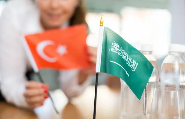 一位积极的年轻女子在桌子上挂着沙特阿拉伯的小旗 旁边放着几瓶水和土耳其的国旗 — 图库照片