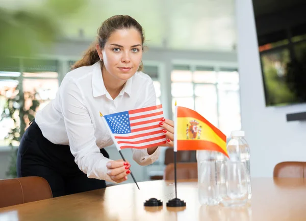 一位积极的年轻女士把美国的小国旗放在西班牙国旗旁边的桌子上 并在会议室里放上几瓶水 — 图库照片