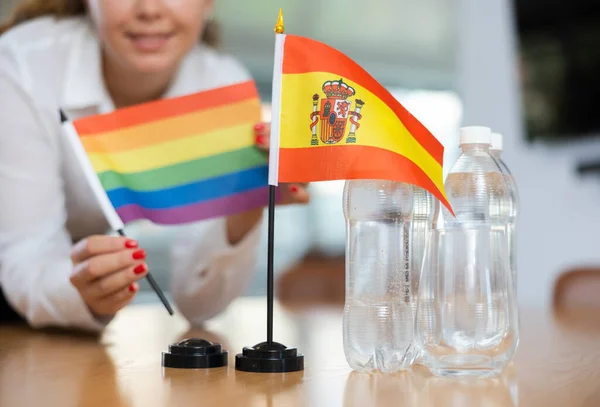会議室で正の若い女性によってそれの隣に置かれた水とLgbtのフラグのボトルとテーブルの上にスペインの小さな旗 — ストック写真