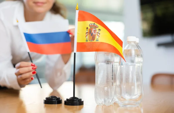 正の若い女性によってそれの横に置かれたロシアの水とフラグのボトルとテーブルの上にスペインの小さな旗 — ストック写真