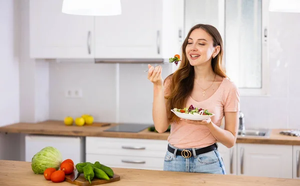 台所のテーブルに立って野菜のサラダを食べるスマイリング菜食主義者の女性 — ストック写真