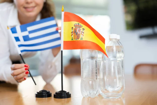 正の若い女性によってそれの横に置かれたギリシャの水とフラグのボトルとテーブルの上にスペインの小さな旗 — ストック写真