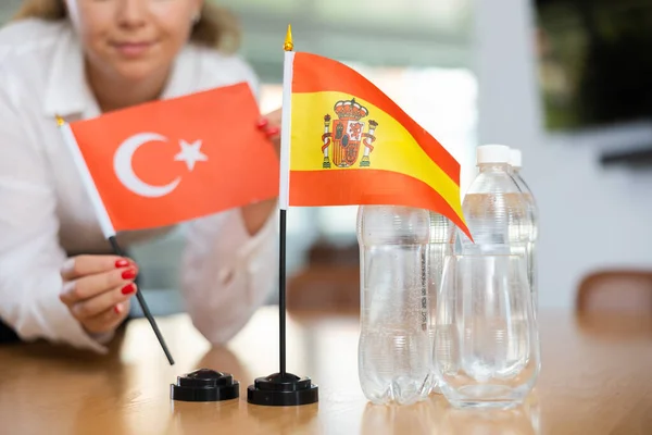 Little flag of Spain on table with bottles of water and flag of Turkey put next to it by positive young woman