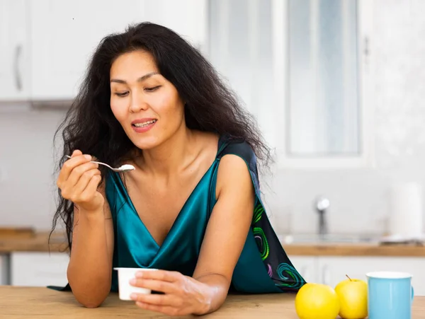 台所のテーブルに立って陽気なアジアの女性とヨーグルトを食べる 朝に乳製品を食べる女性 健康的な朝食 — ストック写真