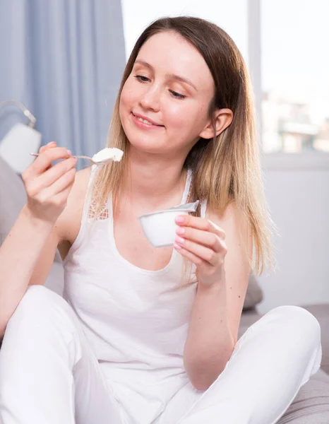 年轻的女人坐在家里的沙发上喝着酸奶 — 图库照片