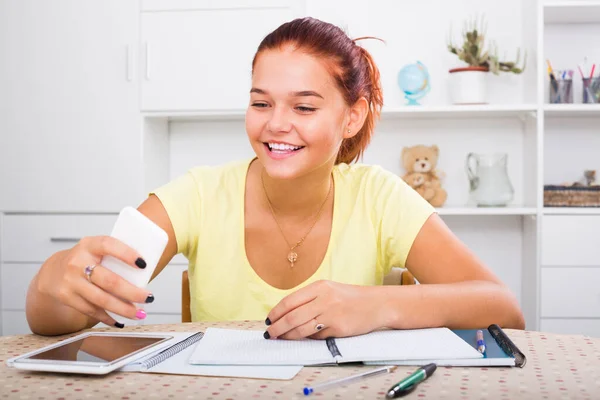 坐在桌边做作业的时候 穿着T恤衫快乐地微笑着的少女在做着自拍 — 图库照片