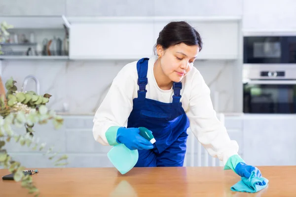 オフィスキッチンで消毒剤で家具にほこりを拭くユニフォームとゴム手袋を身に着けている勤勉な若い女性 熟練した清掃サービス労働者 — ストック写真