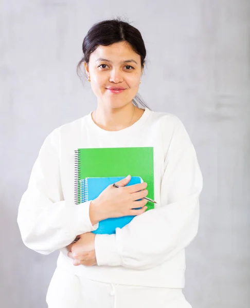 Attractive positive young Asian female college student in white casual clothes holding notebooks standing on gray background. Studying concept..