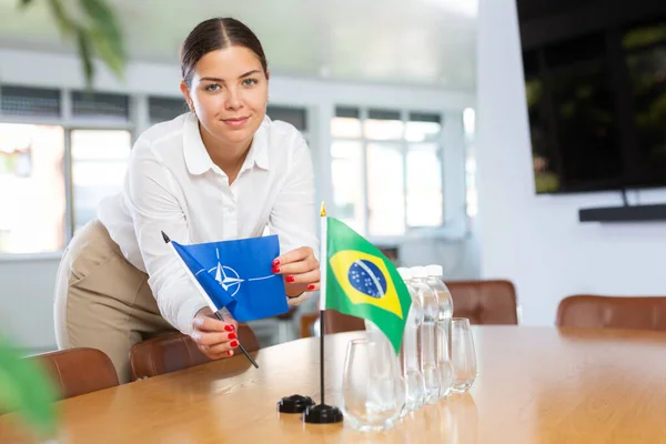 会議室でブラジルの旗と水のボトルの横にテーブルの上にNatoの小さな旗を置く積極的な若い女性 — ストック写真