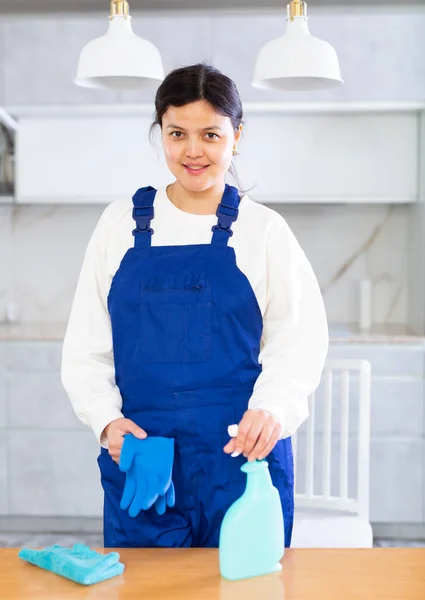 穿着蓝色工作服 面带微笑的年轻清洁女士的画像 她站在家里或办公室厨房轻盈的桌子旁边 手里拿着破布和洗涤剂喷雾 准备清理的女工 — 图库照片