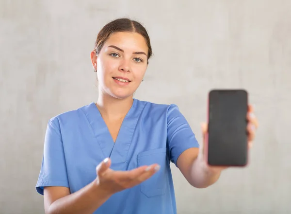 Uśmiechnięta Pielęgniarka Niebieskim Mundurze Poleca Mobilną Aplikację Medyczną Wyciągając Dłoń — Zdjęcie stockowe