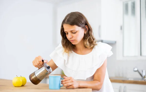 若いです女性は自宅でキッチンテーブルに立って コーヒーを注ぐ — ストック写真
