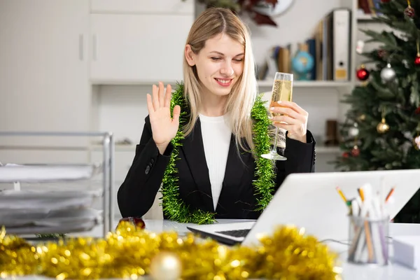 若い女性従業員の笑顔はクリスマスの時間の間にオフィスでラップトップ経由でビデオ通話を持っています シャンパンを飲みます — ストック写真