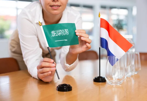 女性秘書は交渉のためのオフィスを準備します 彼女はサウジアラビアとオランダの旗を置きます — ストック写真