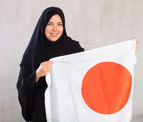 身穿传统黑色头巾的快乐的穆斯林妇女微笑着 举着日本国旗 灰色背景下带有日本国旗的女性穆斯林画像 — 图库照片