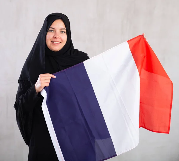 身穿传统黑色头巾的快乐微笑的穆斯林妇女举着法国国旗 灰色背景下带有法国国旗的女性穆斯林画像 — 图库照片