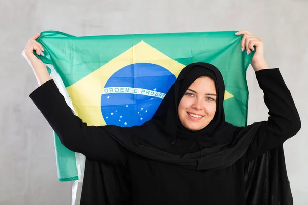 ブラジル国旗を掲げる黒い伝統的なヒジャブを身に着けている陽気な若いイスラム教徒女性 — ストック写真