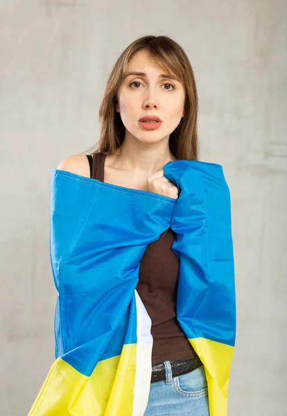 年轻而悲伤的爱国女子 一边用乌克兰国旗包裹着自己 一边看着灰墙背景的相机 — 图库照片