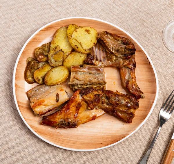 Portion of hearty lunch on plate - pieces of baked rabbit with potato slices. Dish is complemented with glass of red wine