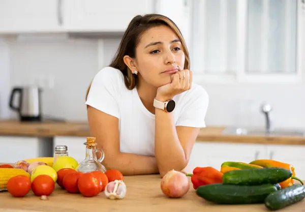 Sad young woman looking at vegetable at kitchen, dieting problems