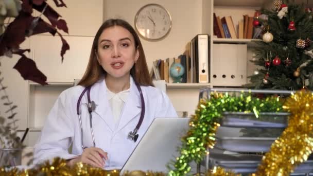 若いですCaucasian医師女性でテーブルに座って装飾されたオフィスでクリスマスとカメラを見て — ストック動画