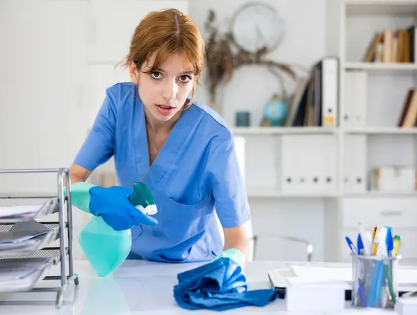 Young woman cleaner in overall polishing table in office with rag and detergent.