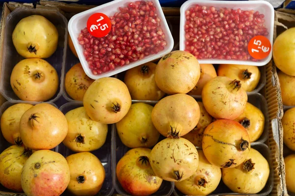 Photo shows fruit shop window with display of pomegranate fruits. Fruits are laid out in plastic containers, customers are also offered already peeled ruby sweet and sour seeds in individual packaging