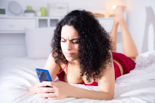 Portrait of young dissatisfied woman in lingerie using phone lying in bed