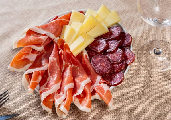 On beige cloth tablecloth - snack plate with meat delicate - sliced sausages and morsels of cheese. Convolute thin slices of ham bacon and complement with circles of butifarra sausage and cheese brie