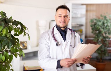 Man doctor standing in office and analyzing documentation in his hands. clipart