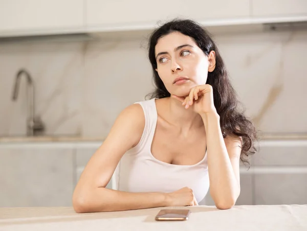 Young upset woman waiting for call on her mobile phone while sitting at table in kitchen