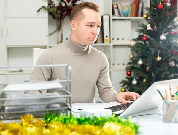 Executive man working in office during Christmastime. Man using laptop in decorated workplace.