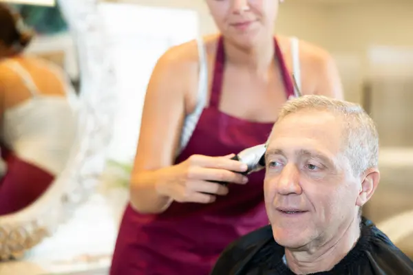Cropped young woman hairdresser performs haircut for elderly man using hair clipper in barbershop. Casual men and women haircuts, hair care, styling, treatment, grooming procedures