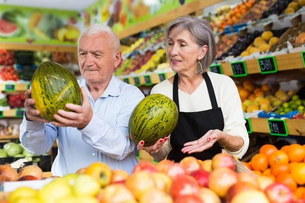 Woman supermarket employee helping mature man to choose watermelon and other fruits