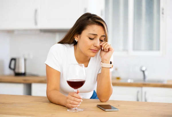 Young depressed Latina sitting alone at home with glass of wine and silent phone feeling abandoned. Upset female suffering from depression after relationship breakup drinking alcohol drink in kitchen
