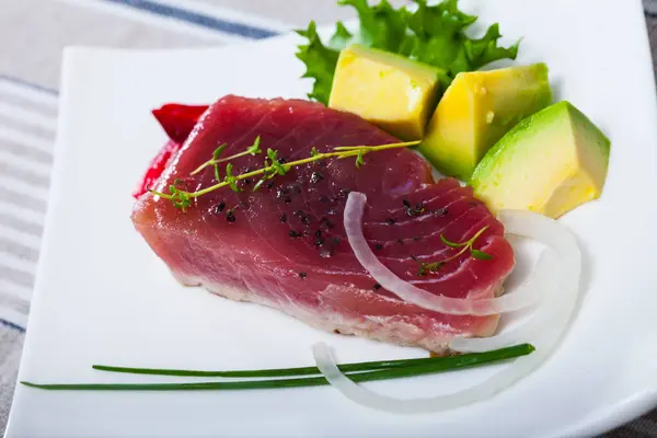 Seafood. Seared tuna steaks sprinkled with sesame with ripe avocado, fresh greens and sun-dried tomatoes on white plate