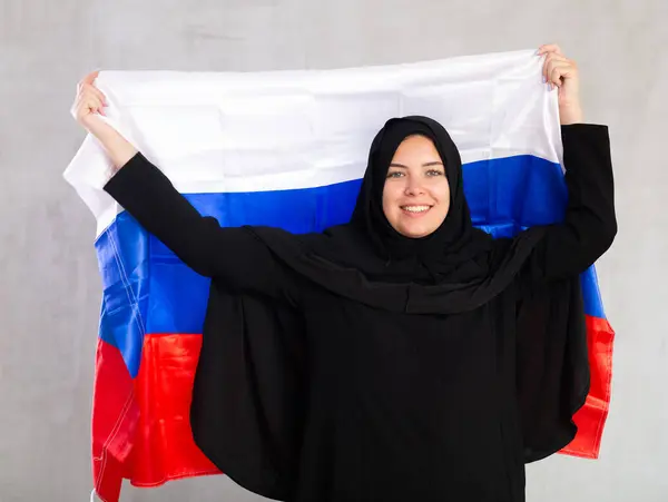 身穿传统黑色头巾的快乐微笑的穆斯林妇女举着俄罗斯国旗 灰色背景下带有俄罗斯国旗的女性穆斯林画像 — 图库照片