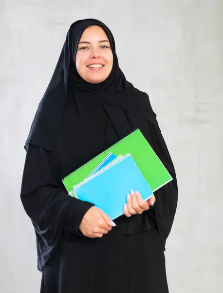 Smiling young female student dressed in traditional islamic clothes holding textbooks