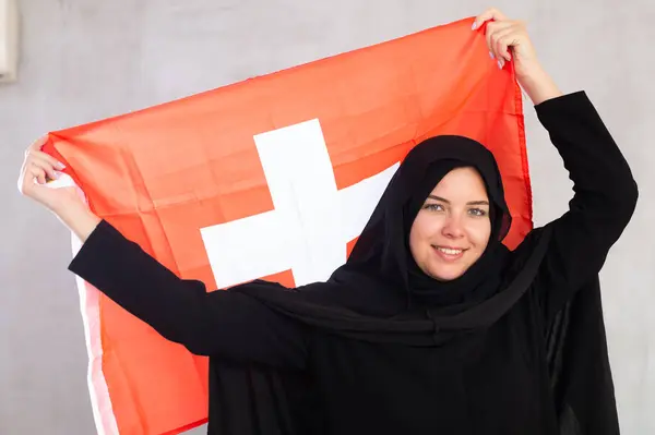 スイスの国旗を掲げる黒い伝統的なヒジャブを身に着けている陽気な若いイスラム教徒の女性 — ストック写真