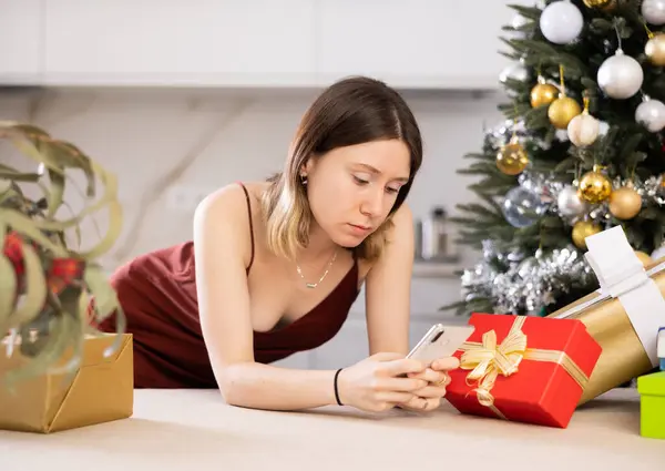Frustrated young woman at home against the background of a Christmas tree with presents, is typing a message to someone on .her mobile phone
