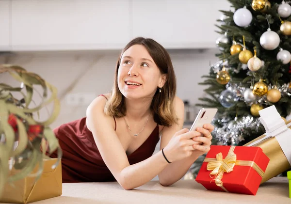 Positive young woman is texting someone on mobile phone while at home against the background of a Christmas tree with presents