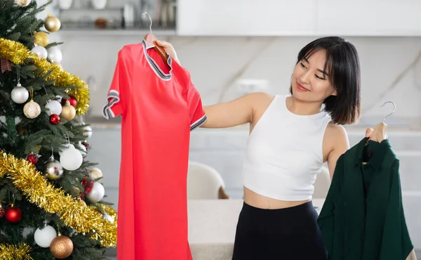 年轻的亚洲女人看到手中拿着可供选择的衣服 站在自家圣诞树旁 感到很高兴 但并不满意 — 图库照片