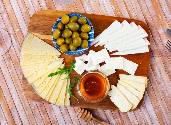 在木制桌子上有切菜板 上面有各种奶酪 腌制橄榄 轻质酒精饮料的精美小吃 奶酪切片用牛油果叶装饰 — 图库照片