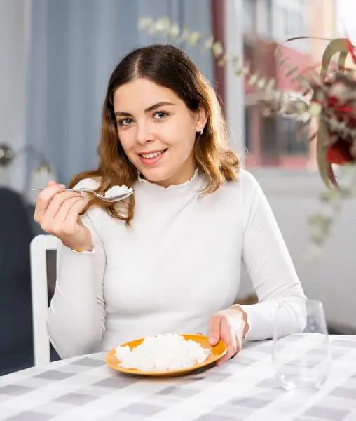 ポジティブな若い女性は アパートのテーブルに座っている間 食事と沸騰米を食べています — ストック写真