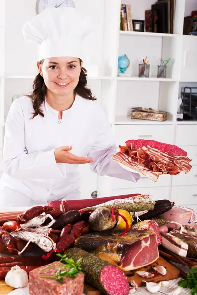 Cook woman costs near table with smoked products and sausage and holds in hand plate with jamon
