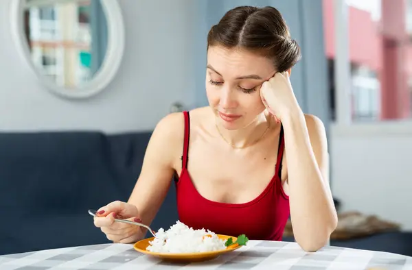 Young sad woman eating rice while sitting at the table in the room. Diet concept and diet foods