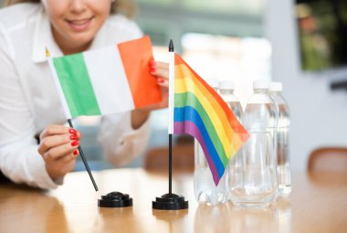 Unrecognizable woman preparing room for international negotiations and communication discussions of leaders. Lady sets miniatures flags of LGBT and Italy on table. Unfocused shot clipart